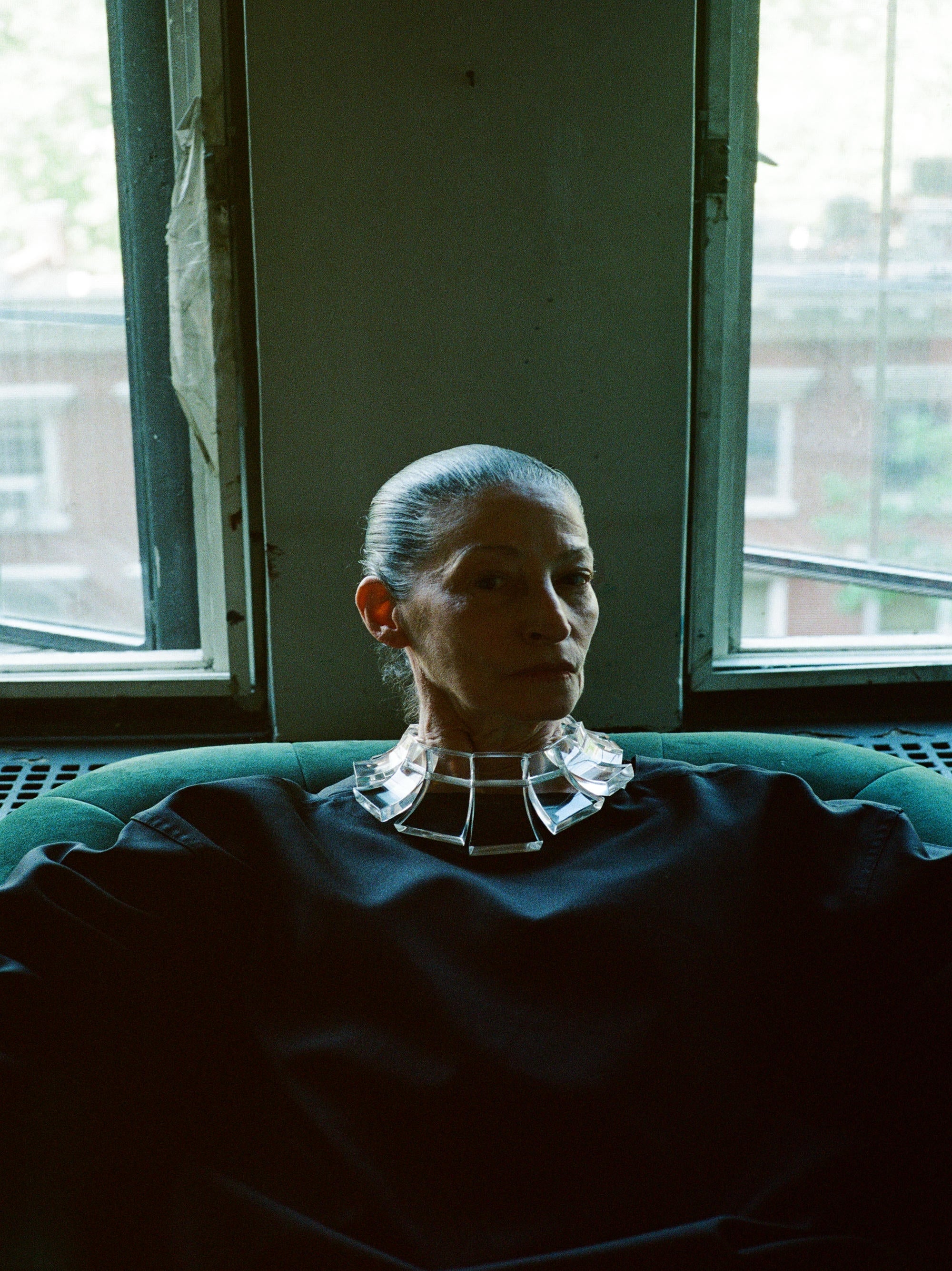 Person sitting on a couch in a dimly lit room with large windows wearing an acrylic necklace