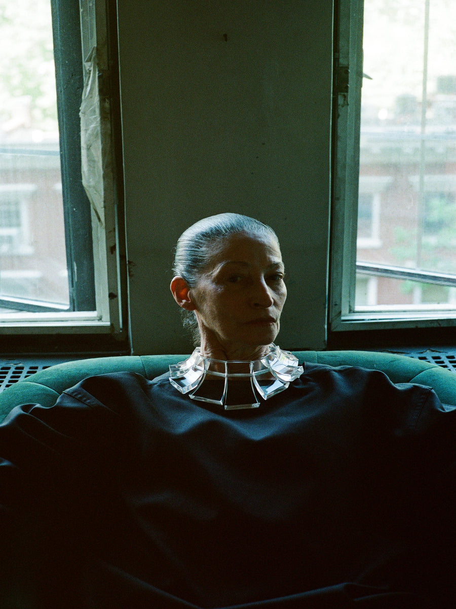 Person sitting on a couch in a dimly lit room with large windows wearing an acrylic necklace
