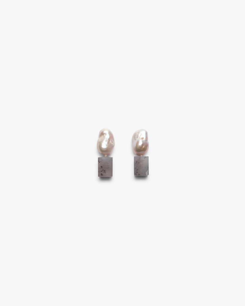 A pair of minimalist drop earrings featuring organic baroque pearls suspended from square, textured gray stone pendants, set against a clean white background.