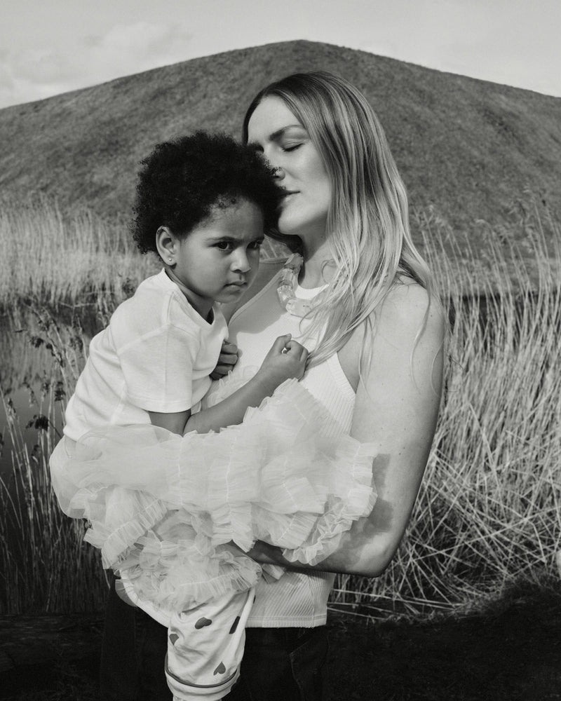 A woman holding a young child with a mountain crystal necklace around their necks, in a natural outdoor setting.