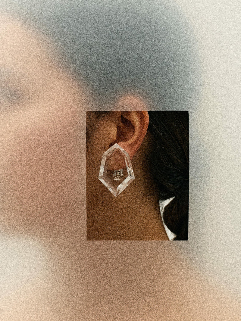 a blurry image of a person wearing A pair of transparent acrylic earrings with a geometric shape, attached to silver plated brass ear clips.