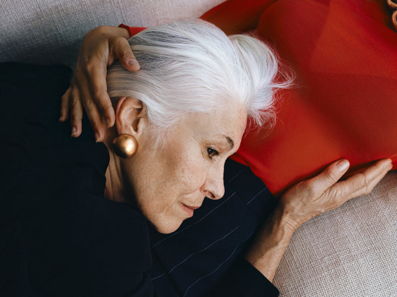 Woman with white hair and gold earrings sitting on a red cushion.