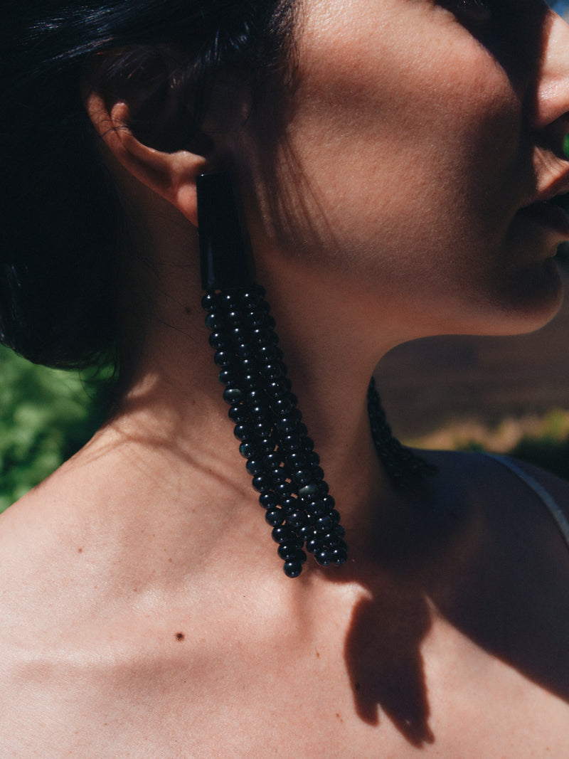 Close-up of a person wearing a long, dark beaded earring with a blurred natural background.