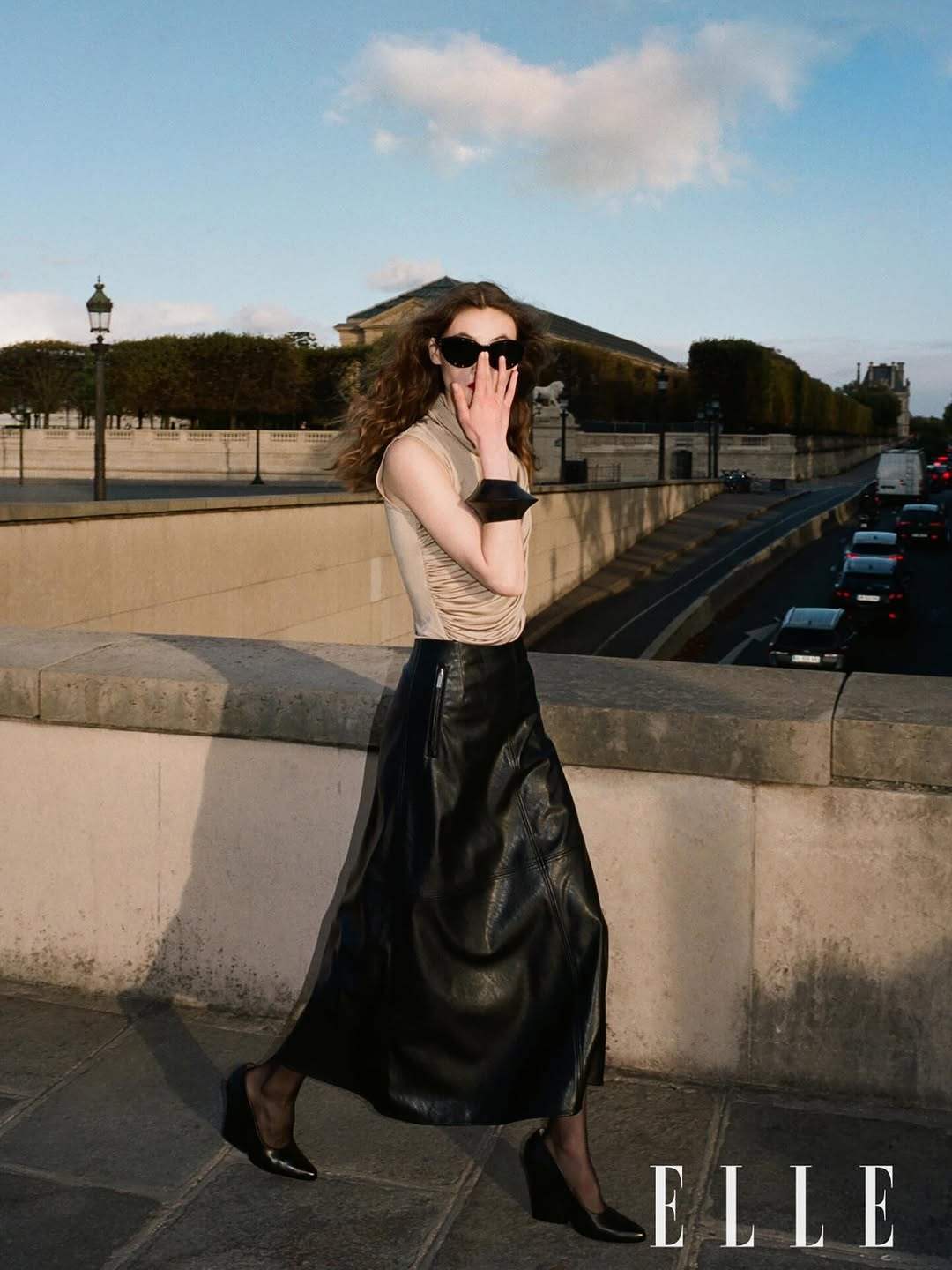 Woman in sunglasses and a black leather skirt standing on a bridge with a cityscape background, featuring the 'ELLE' magazine logo.