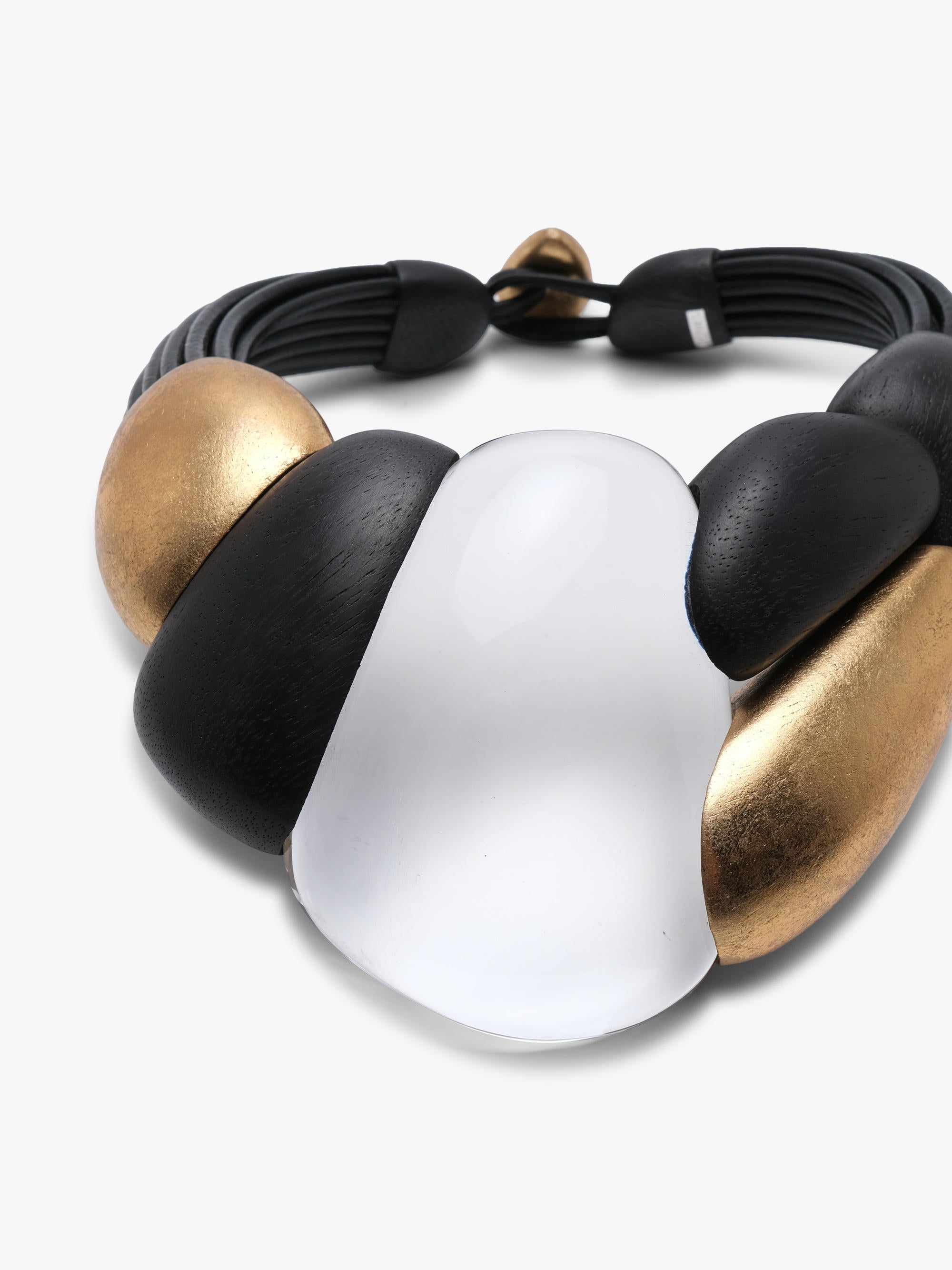A modern statement collar necklace featuring a black leather cord base with sculptural matte black and gold beads, a polished transparent oval centerpiece, and a small gold clasp, shown on a light gray background.