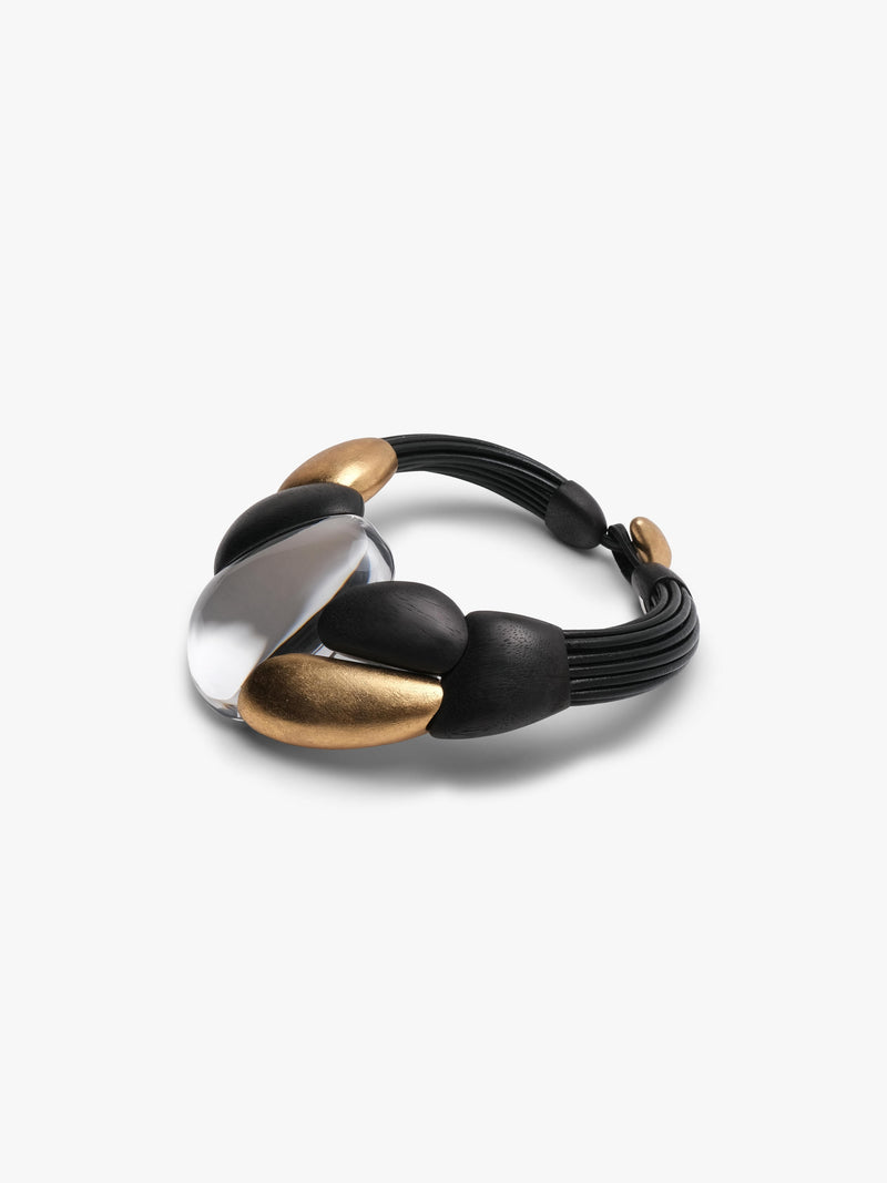 A modern statement collar necklace featuring a black leather cord base with sculptural matte black and gold beads, a polished transparent oval centerpiece, and a small gold clasp, shown on a light gray background.