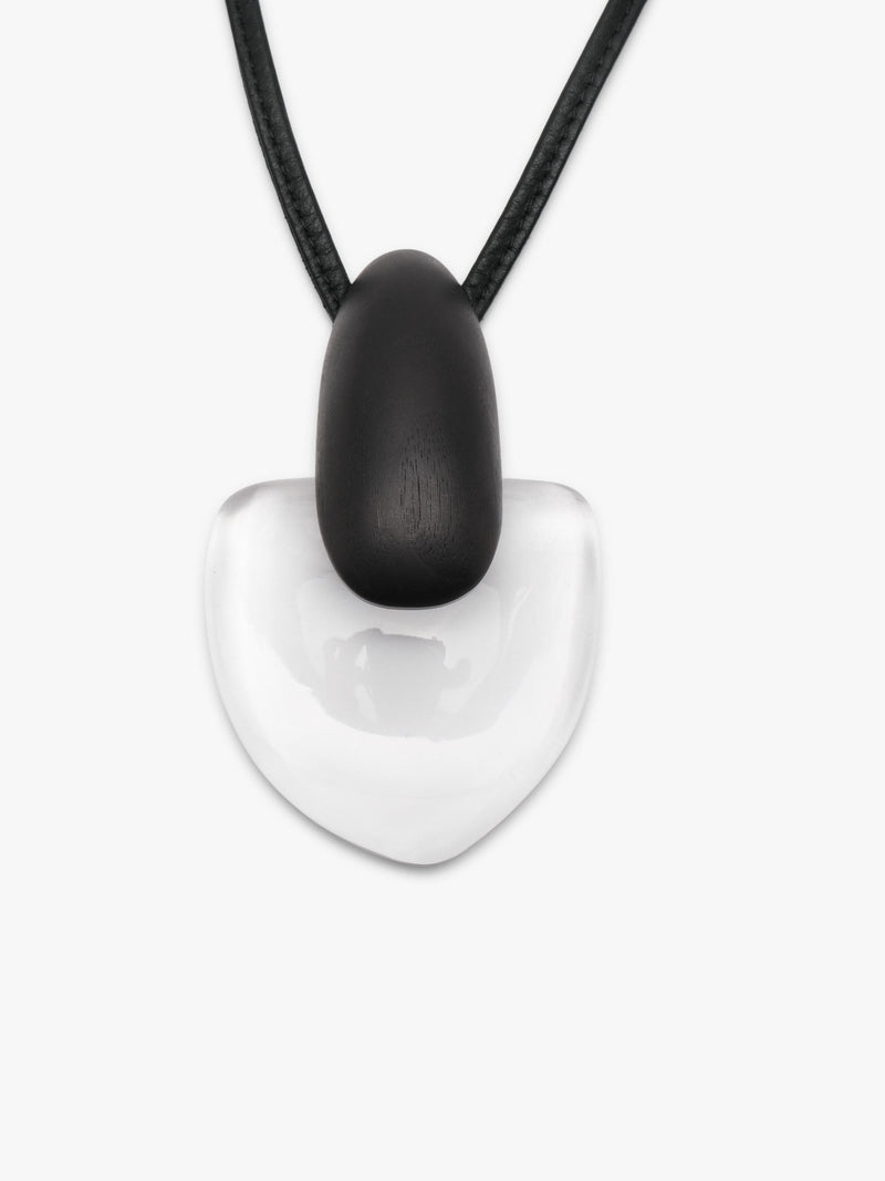 A pendant featuring a smooth, teardrop-shaped black pendant resting on a larger, polished transparent heart-shaped stone, suspended from a black leather cord, shown against a plain light gray background.