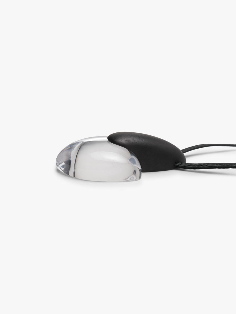 A pendant featuring a smooth, teardrop-shaped black pendant resting on a larger, polished transparent heart-shaped stone, suspended from a black leather cord, shown against a plain light gray background.