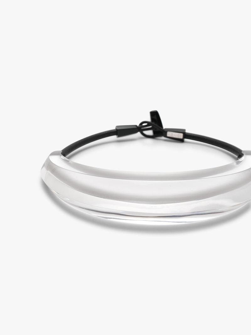 A minimalist crescent-shaped collar necklace featuring a wide, semi-transparent curved band with a black leather cord and toggle clasp, shown on a plain light gray background