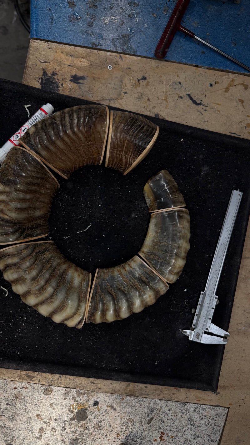 A curved horn or antler-like decorative piece with layered brown and tan ridges, laid on a black mat with calipers and a marker nearby on a workshop table.