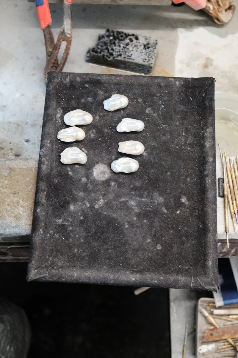Six small white irregular baroque pearls arranged on a dark rectangular tray or board, photographed in a workshop setting with tools and materials visible around it.