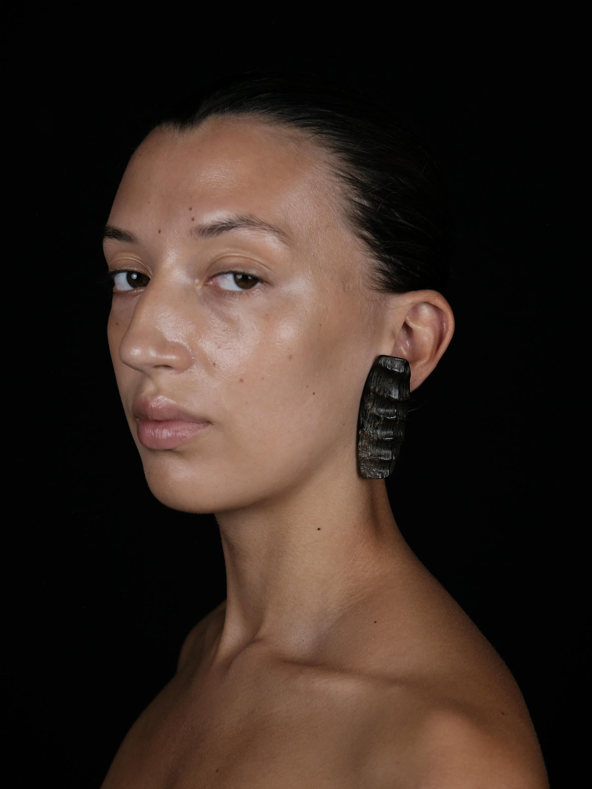 A close-up portrait of a woman with dark hair pulled back, wearing a large textured black earring, set against a solid black background.