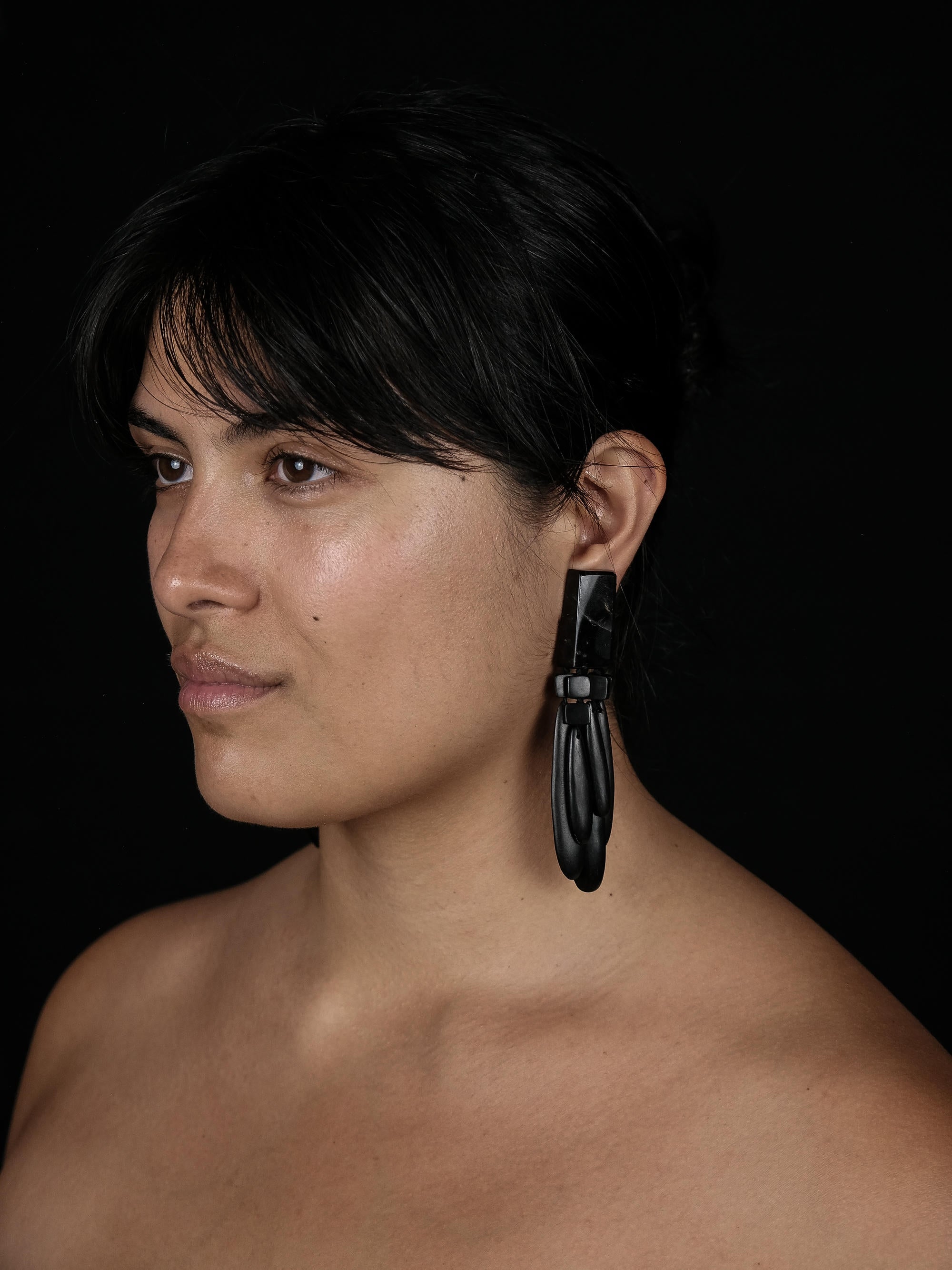 A close-up portrait of a woman with dark hair wearing a large, black, geometric tribal-style dangle earring against a solid black background.