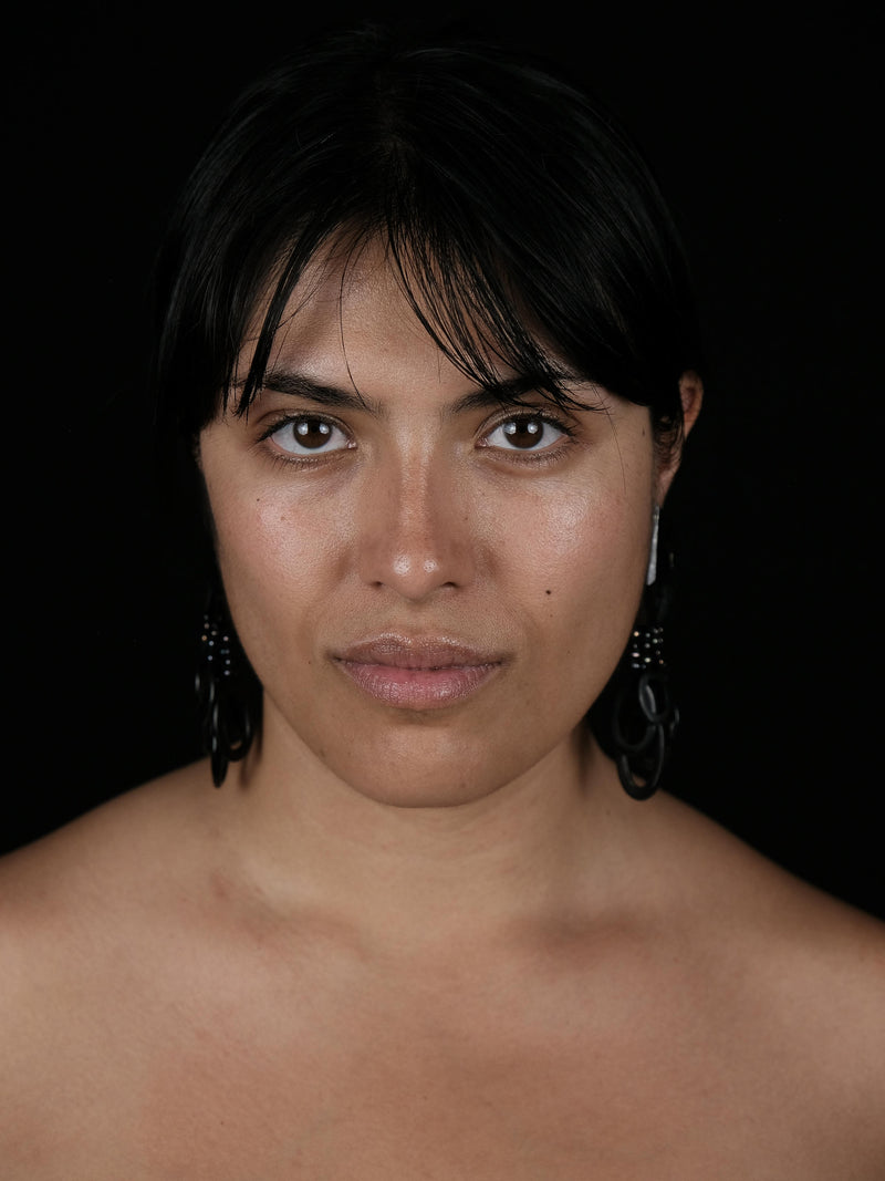 A close-up portrait of a woman with dark hair wearing a large, black, geometric dangling earring against a solid black background.
