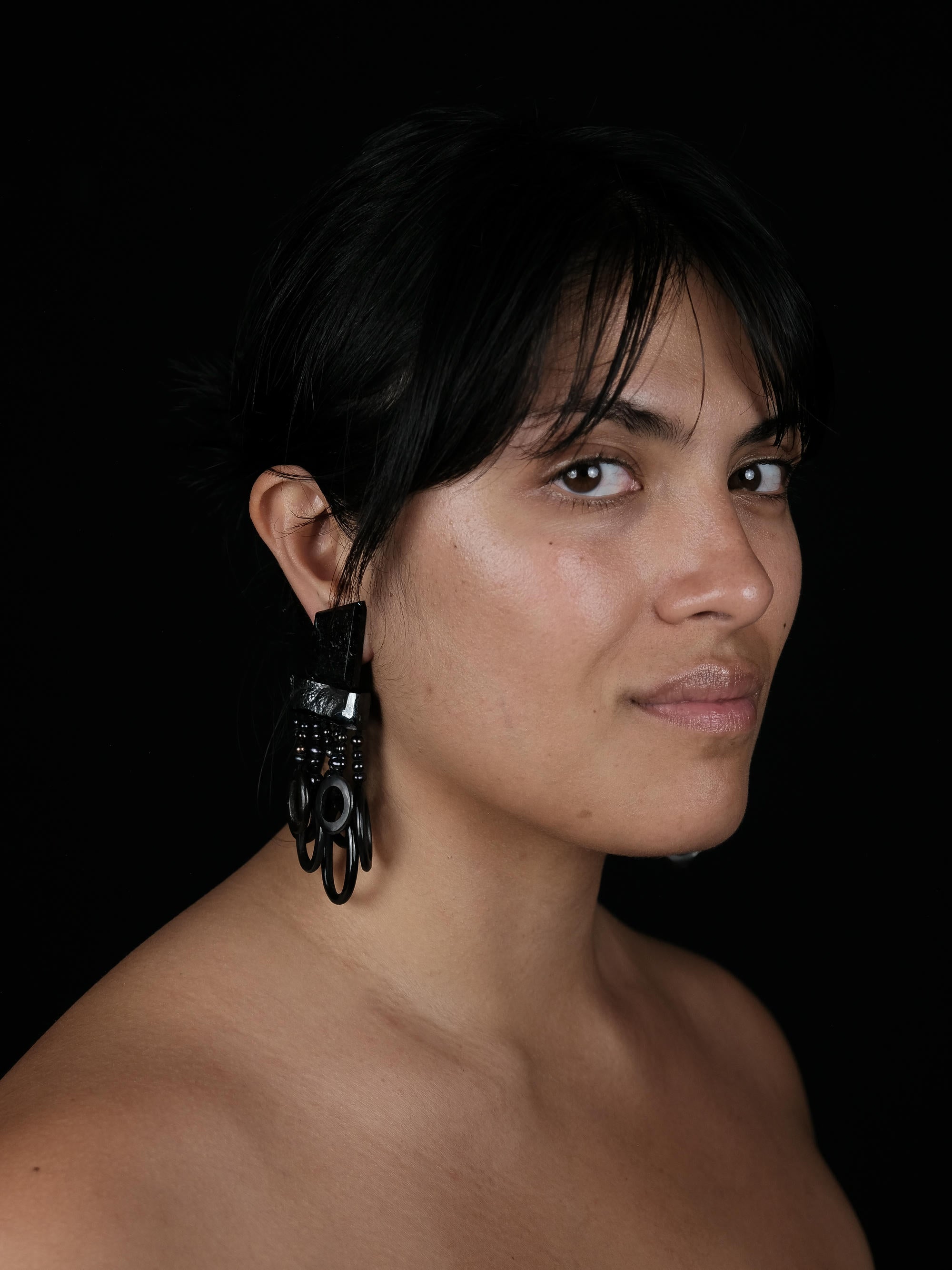 A close-up portrait of a woman with dark hair wearing a large, black, geometric dangling earring against a solid black background.