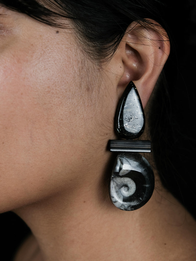 A close-up side profile of a woman wearing a pair of dramatic teardrop-shaped earrings made from black and white marbled stone, set against a solid black background.