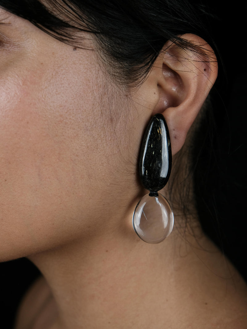 A close-up of a person's ear wearing a teardrop-shaped drop earring with a glossy black top section and a clear glass bottom drop, set against a dark background.