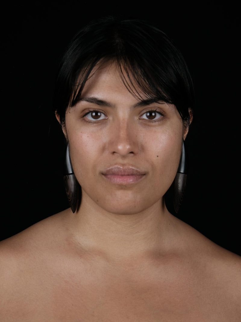 A close-up portrait of a woman with dark hair and large brown and black earrings against a solid black background.
