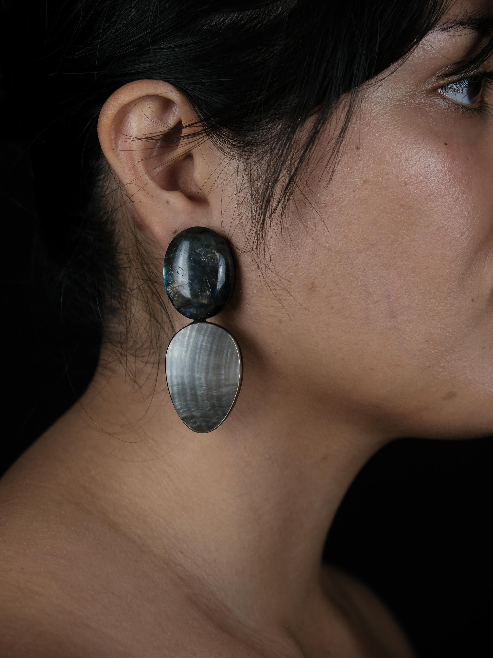 A close-up side profile of a woman wearing a large double-drop statement earring. The earring features a dark, polished oval top stone and a larger, horizontally striped teardrop-shaped bottom stone.