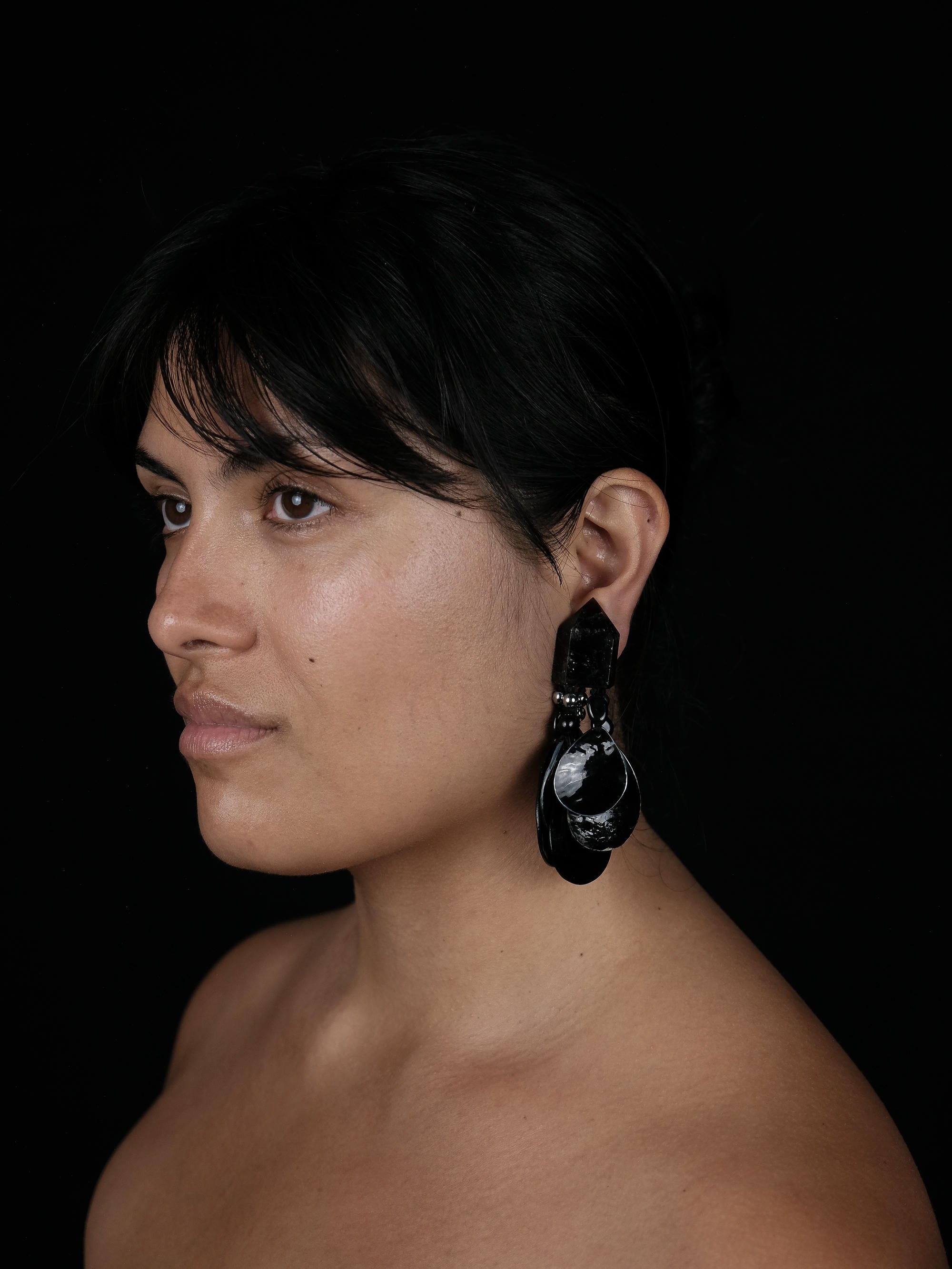 A close-up side profile of a woman with dark hair wearing a large, dramatic black and white sculptural dangle earring against a solid black background.