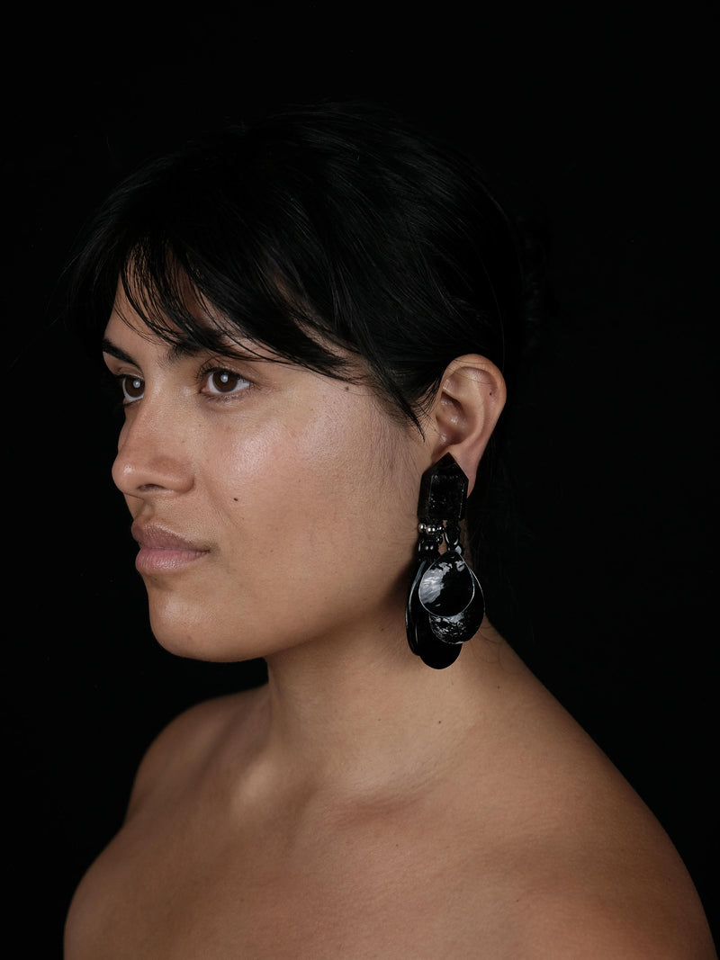 A close-up side profile of a woman with dark hair wearing a large, dramatic black and white sculptural dangle earring against a solid black background.