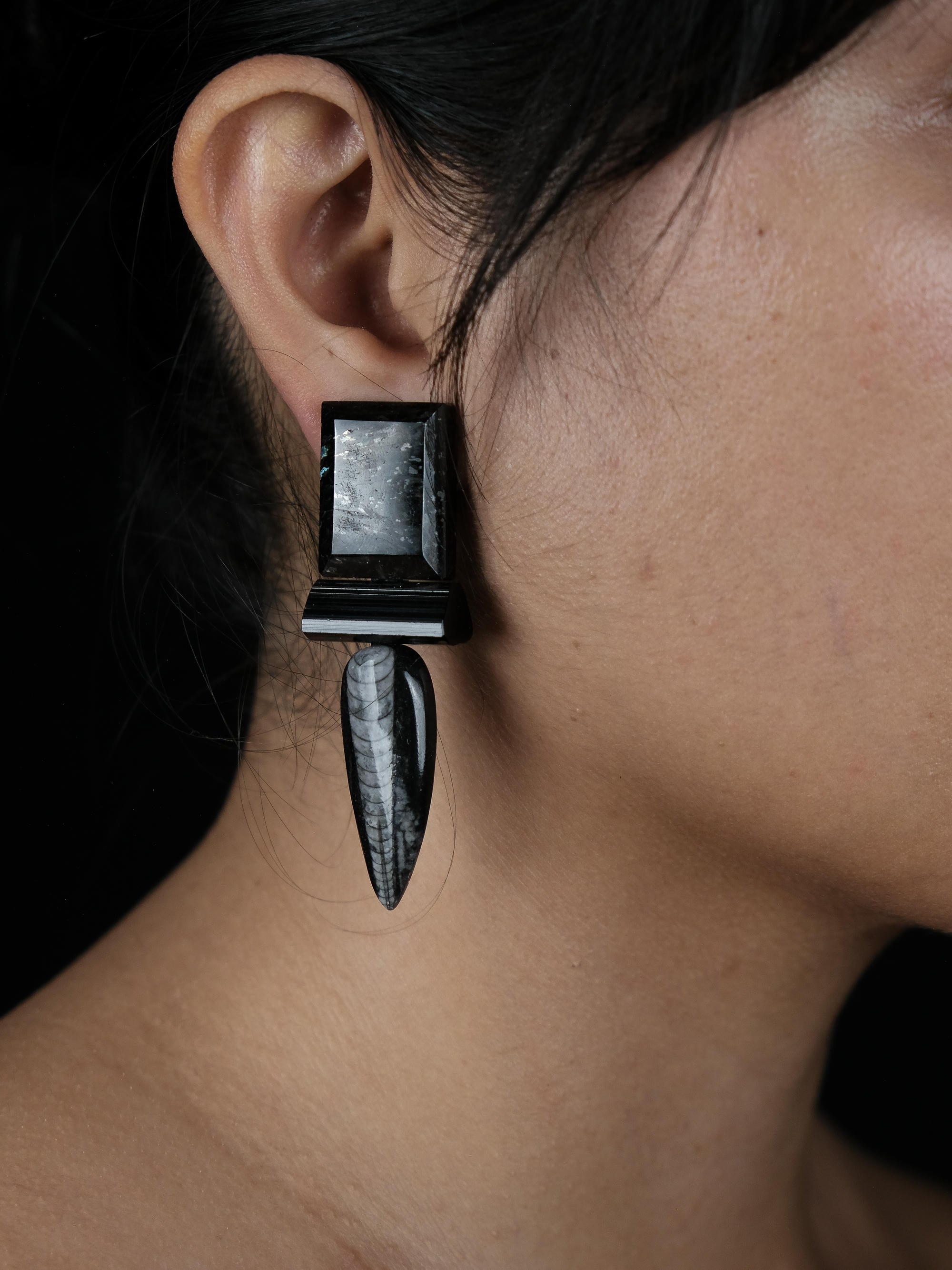 A close-up of a person's ear wearing a sculptural statement earring. The earring features a square black top section with a textured gray inlay, connected to a long, tapered black drop element with a white marbled pattern, set against a dark background.