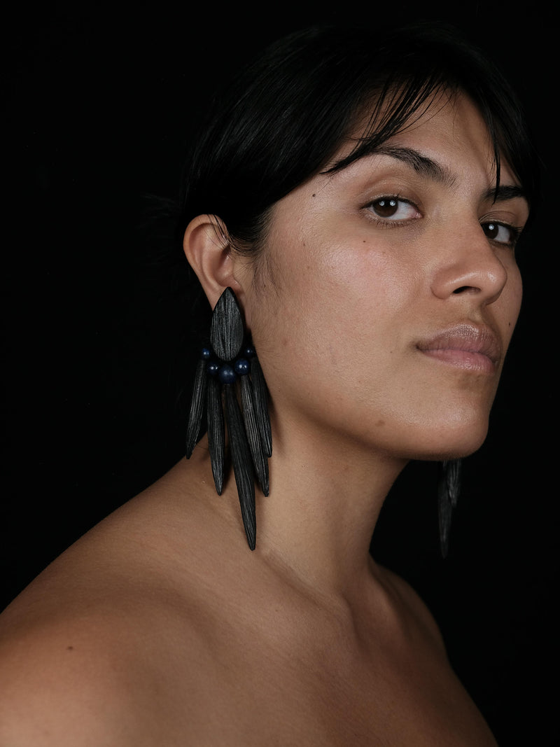 A woman in side profile, wearing large dangle earrings in textured black material, against a black background