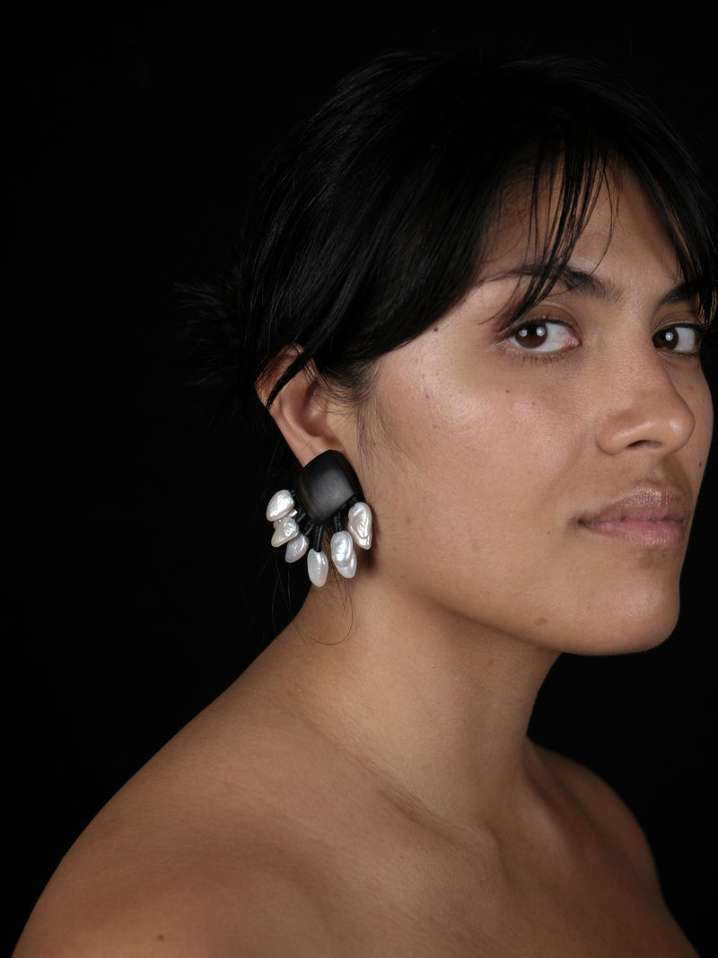 A close-up portrait of a woman with dark hair wearing a dramatic black and white sculptural dangle earring against a solid black background.