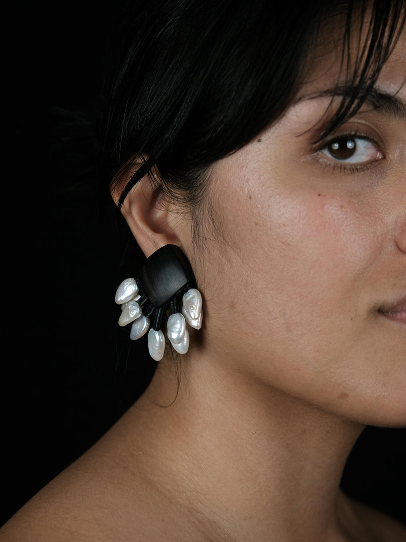 A close-up portrait of a woman with dark hair wearing a dramatic black and white sculptural dangle earring against a solid black background.