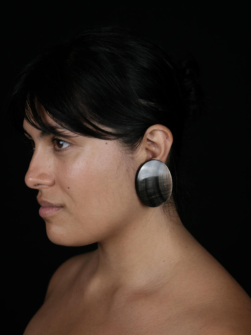 A close-up side profile of a woman with dark hair wearing a large, circular, polished black and white stone earring against a solid black background.