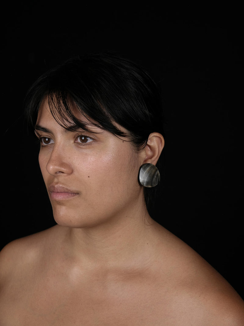A close-up portrait of a woman with dark hair wearing a large, round, silver shelllike earring against a solid black background.