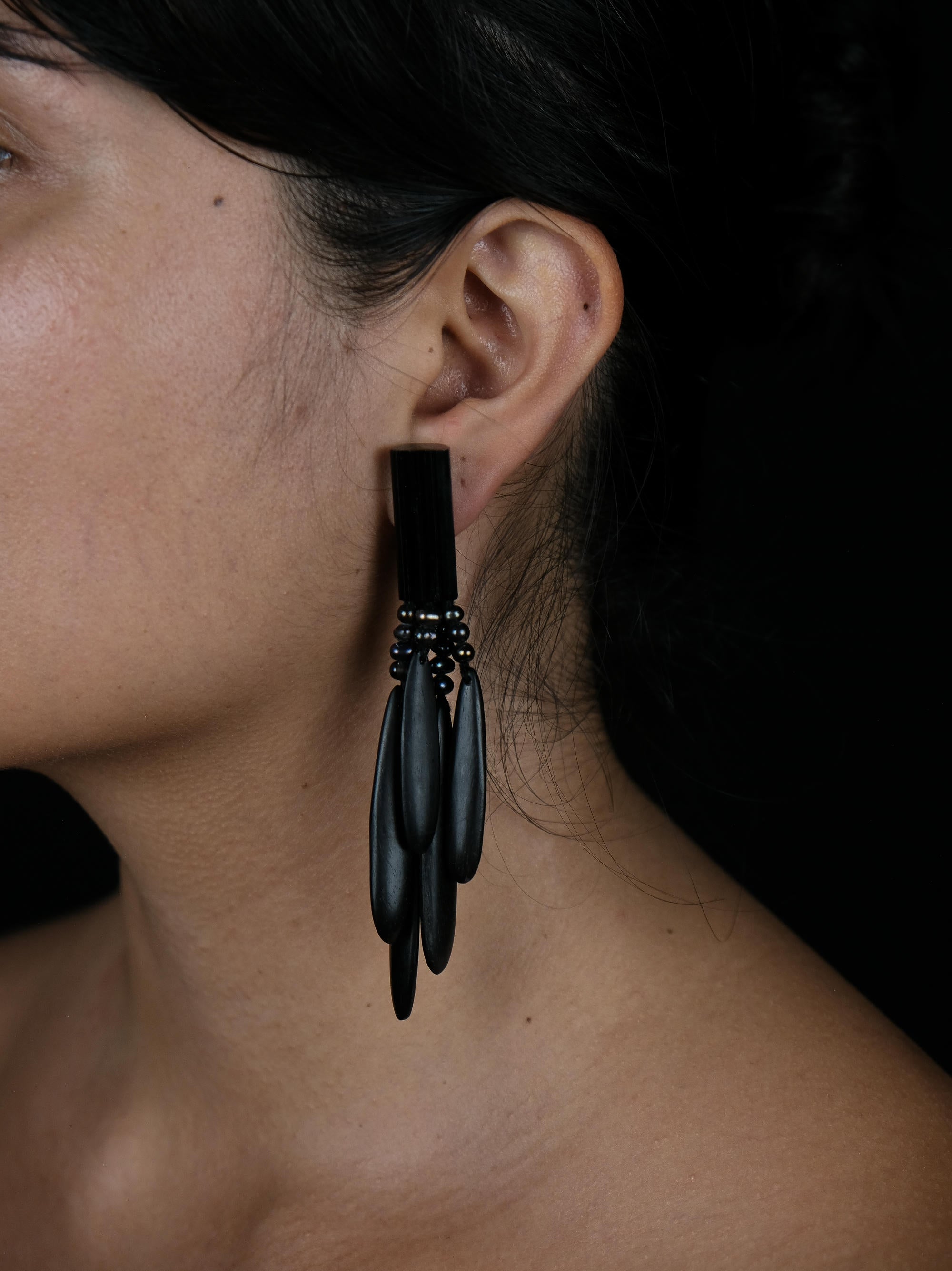 A close-up side profile of a woman wearing a dramatic black statement earring with multiple dangling teardrop-shaped elements and small dark beads, set against a dark background.