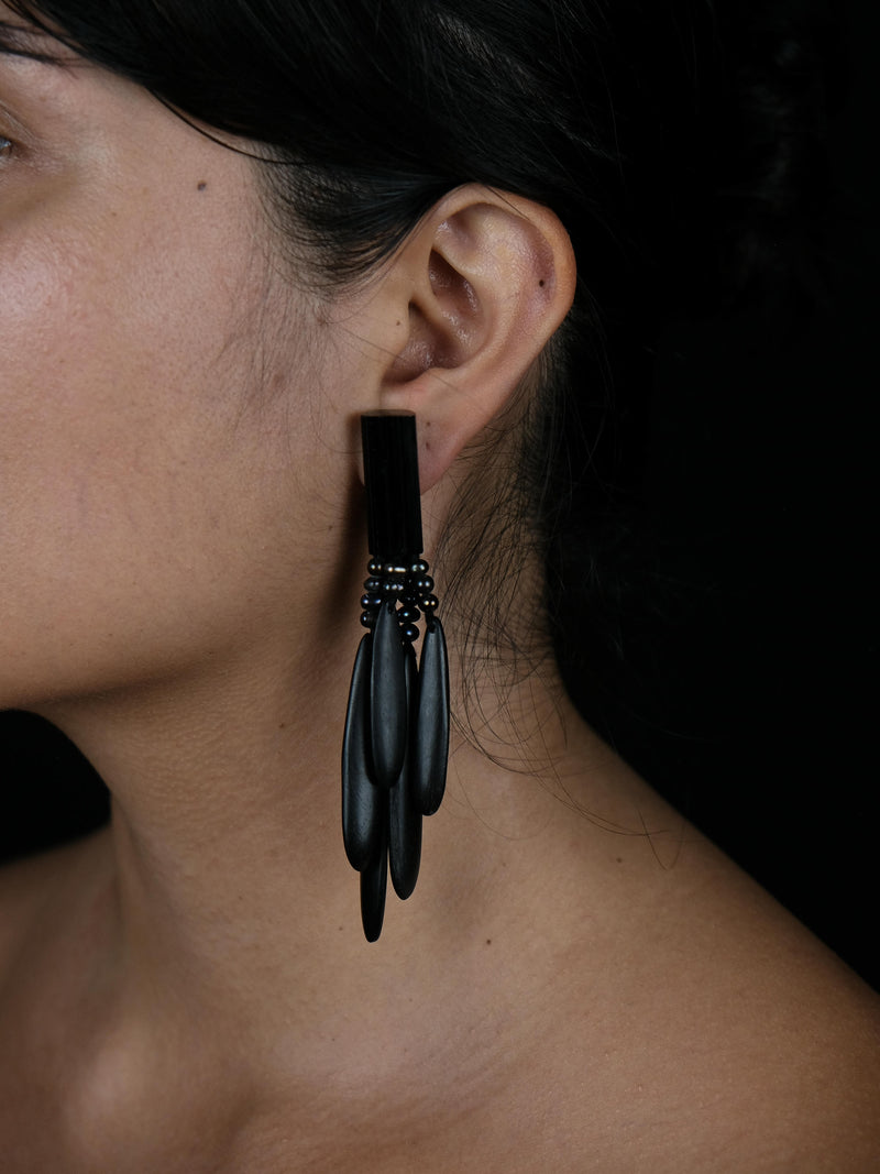 A close-up side profile of a woman wearing a dramatic black statement earring with multiple dangling teardrop-shaped elements and small dark beads, set against a dark background.
