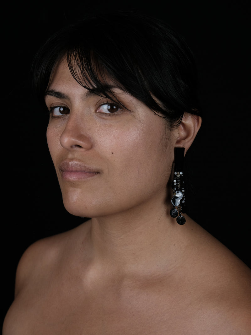 A close-up portrait of a woman with dark hair and bare shoulders, wearing a long dangling earring with black and silver elements, set against a solid black background.