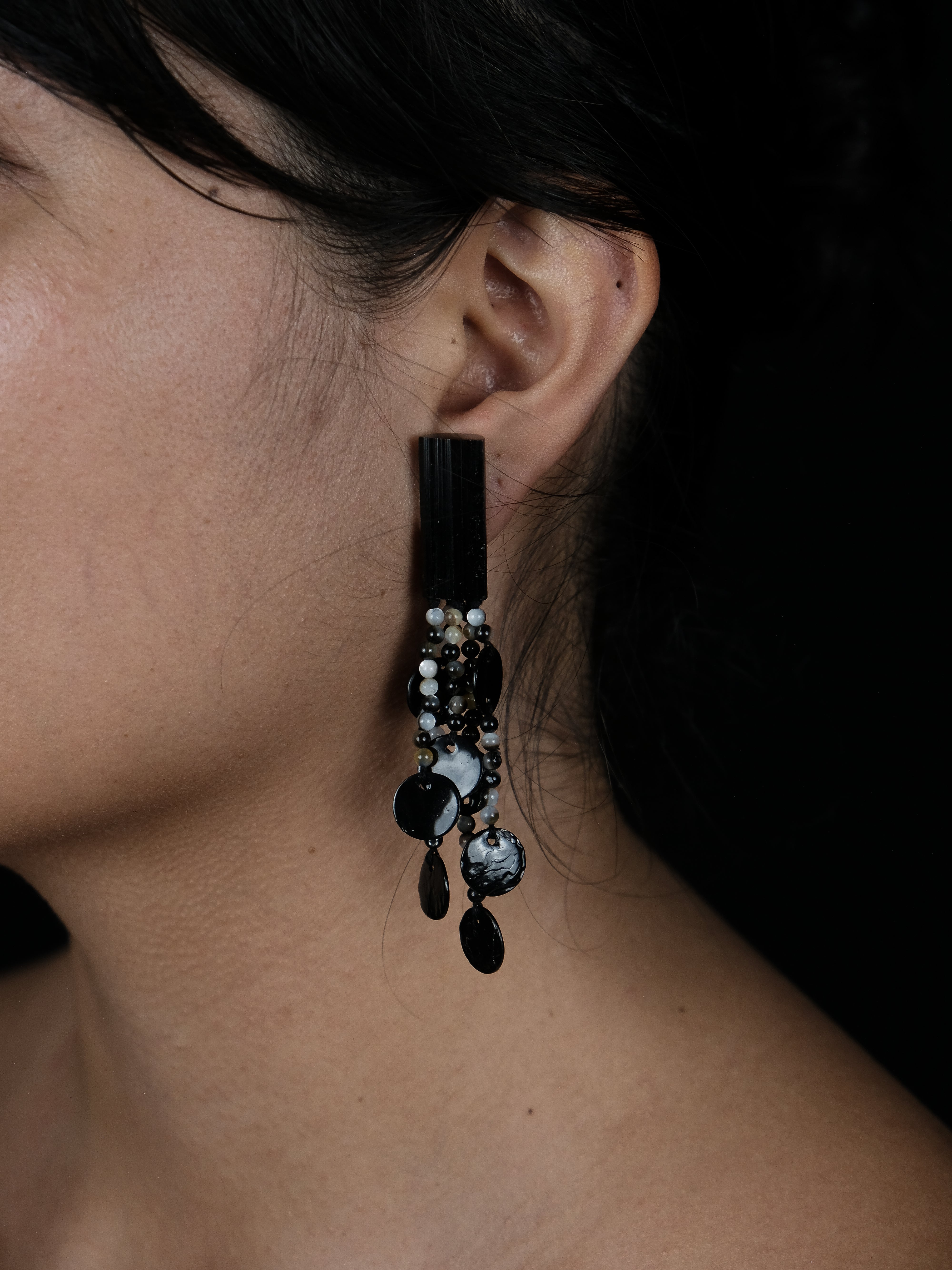 A close-up portrait of a woman with dark hair and bare shoulders, wearing a long dangling earring with black and silver elements, set against a solid black background.