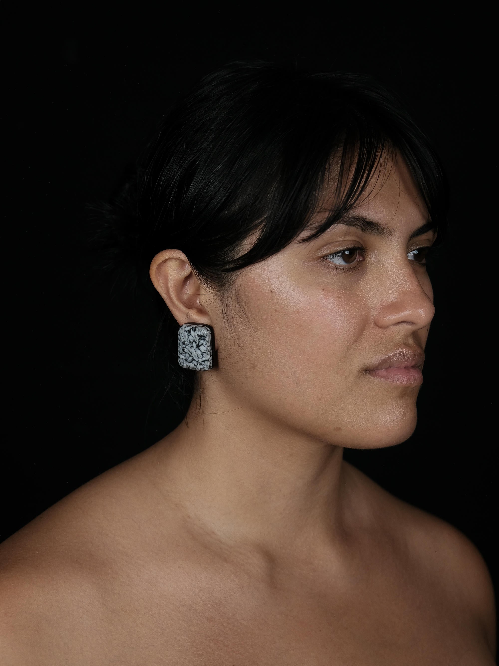 A close-up portrait of a woman in profile wearing a textured square earring against a solid black background.