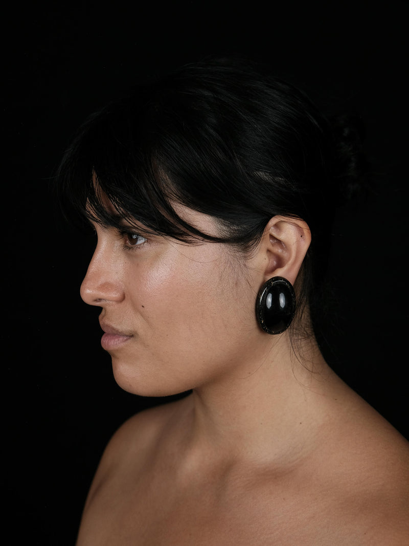 A close-up side profile of a woman with dark hair wearing a large, glossy black oval earring against a solid black background.