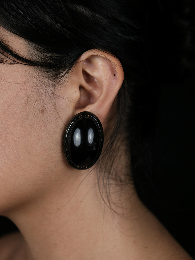 A close-up side profile of a woman with dark hair wearing a large, glossy black oval earring against a solid black background.