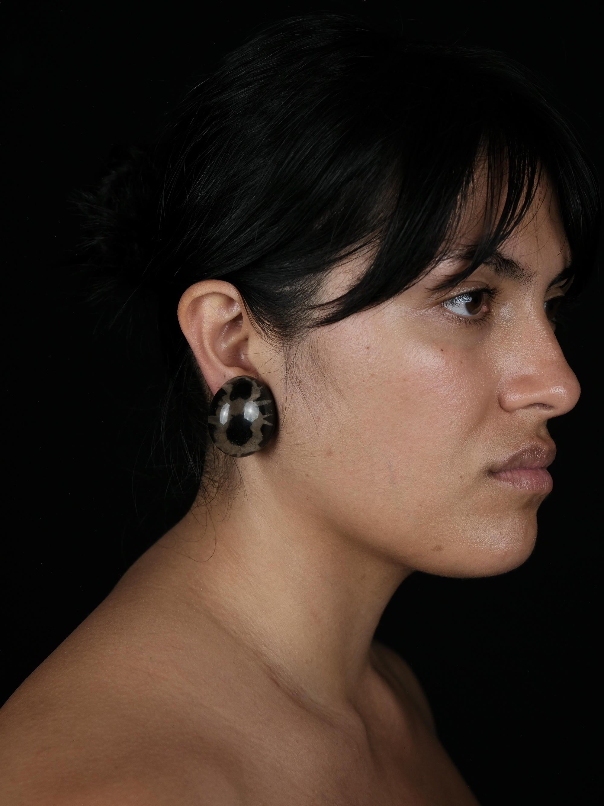 A close-up side profile of a woman with dark hair wearing a large, round, marbled black and brown earring against a solid black background.