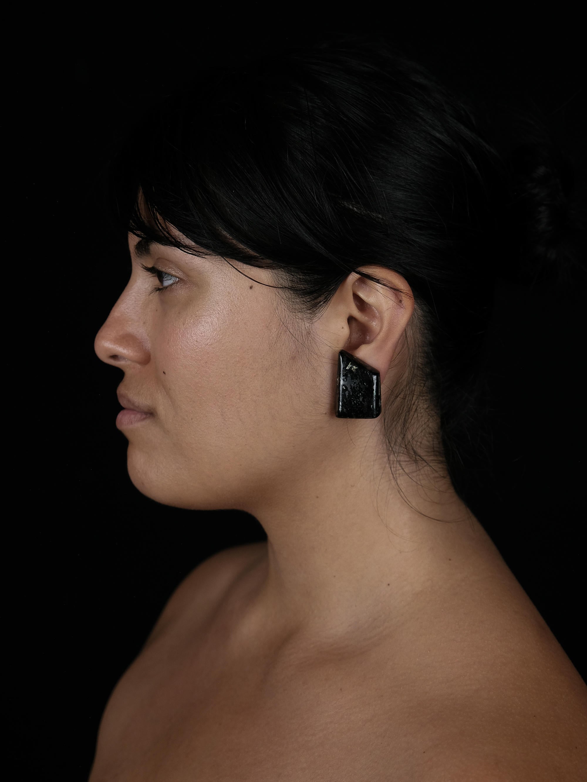 An image of a woman with dark hair, wearing a black earring against a black background