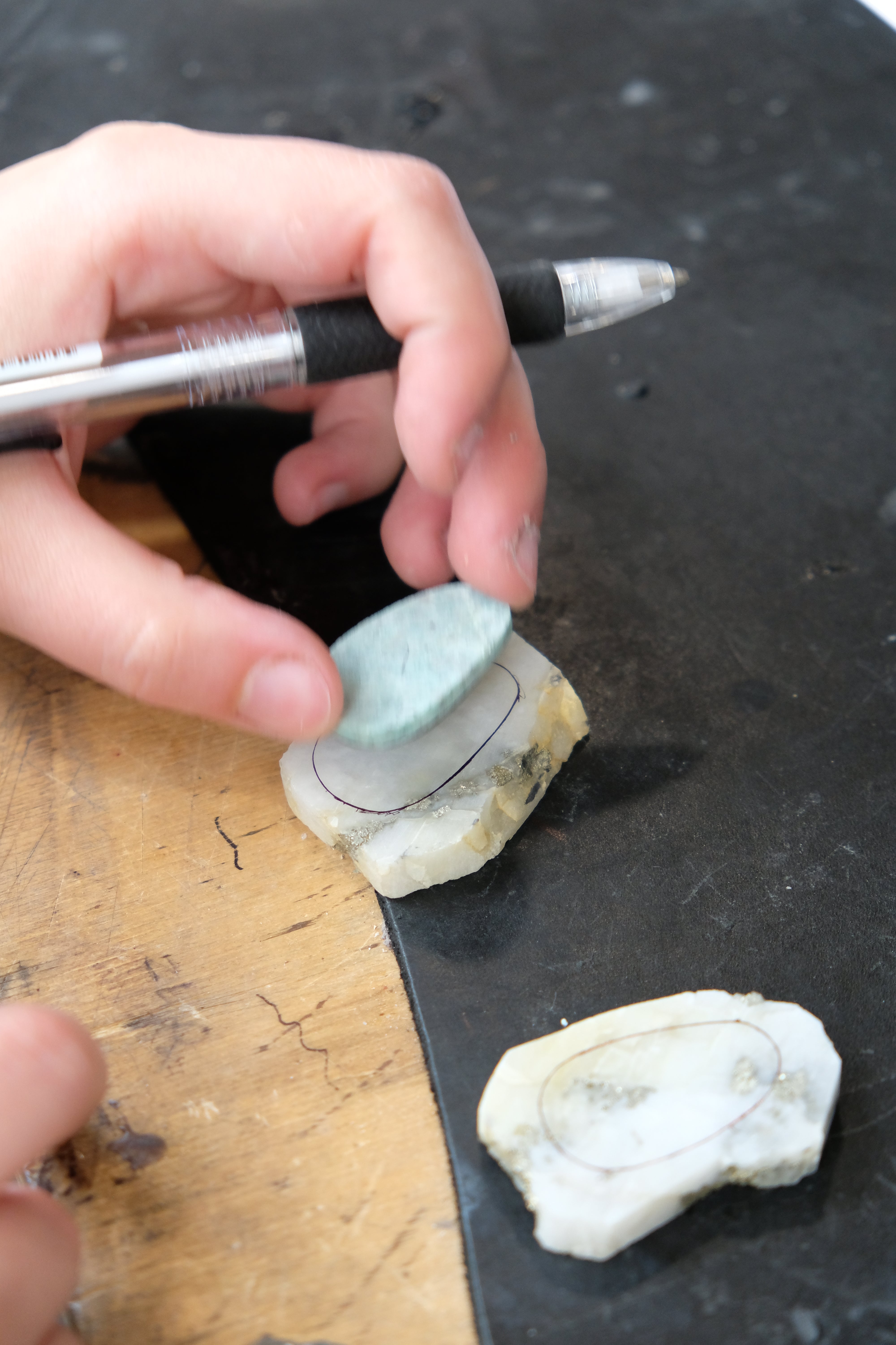 A close-up of a hand holding a small oval-shaped light blue stone or tile over a rough white stone sample marked with a black outline, with a silver pen resting nearby on a dark surface.