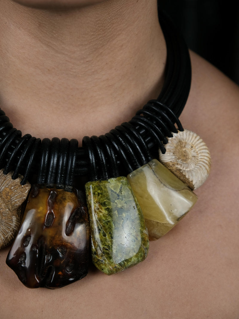 A close up of a woman with black hair, wearing a black cord necklace with large charms in different stones, including amber and green stones, against a black background