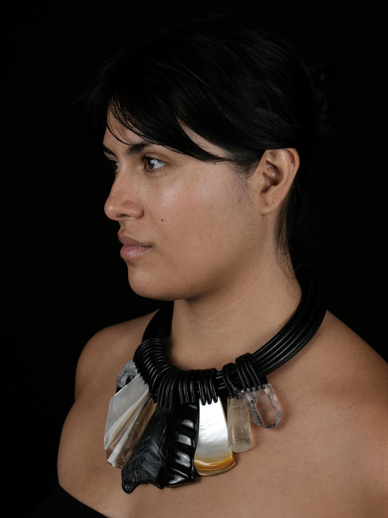 A woman with dark hair, wearing a black leather necklace, with large stonependants in different colours and textures, against a black background