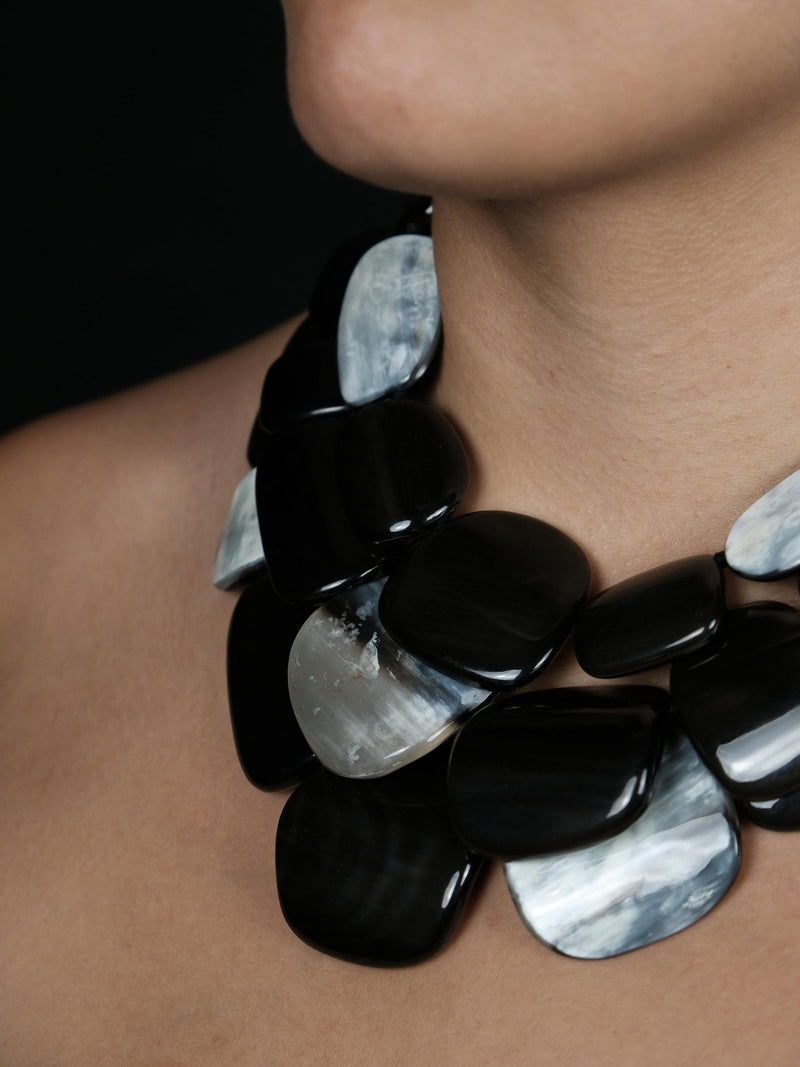 A close-up of a model wearing a chunky statement necklace made of glossy black and marbled silver-gray stone-like pieces.