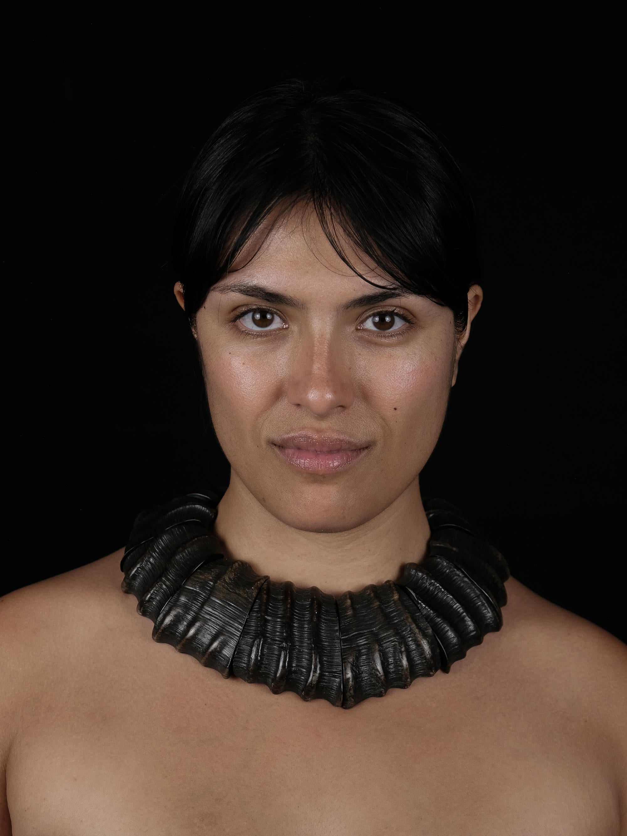 A woman with black hair wearing a large textured necklace in dark brown, against a black background