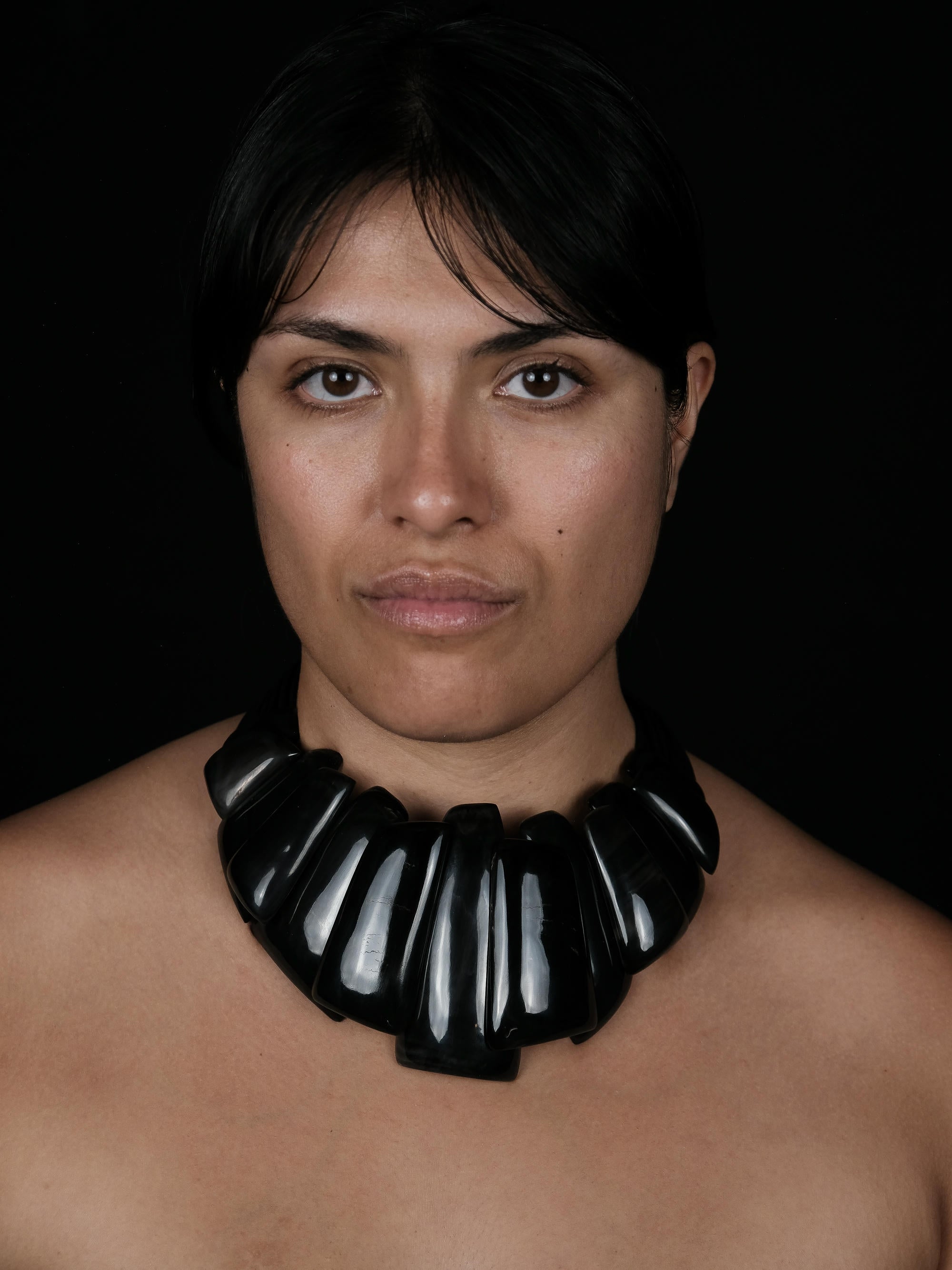 A close-up portrait of a woman wearing a chunky, glossy black collar necklace against a solid black background.
