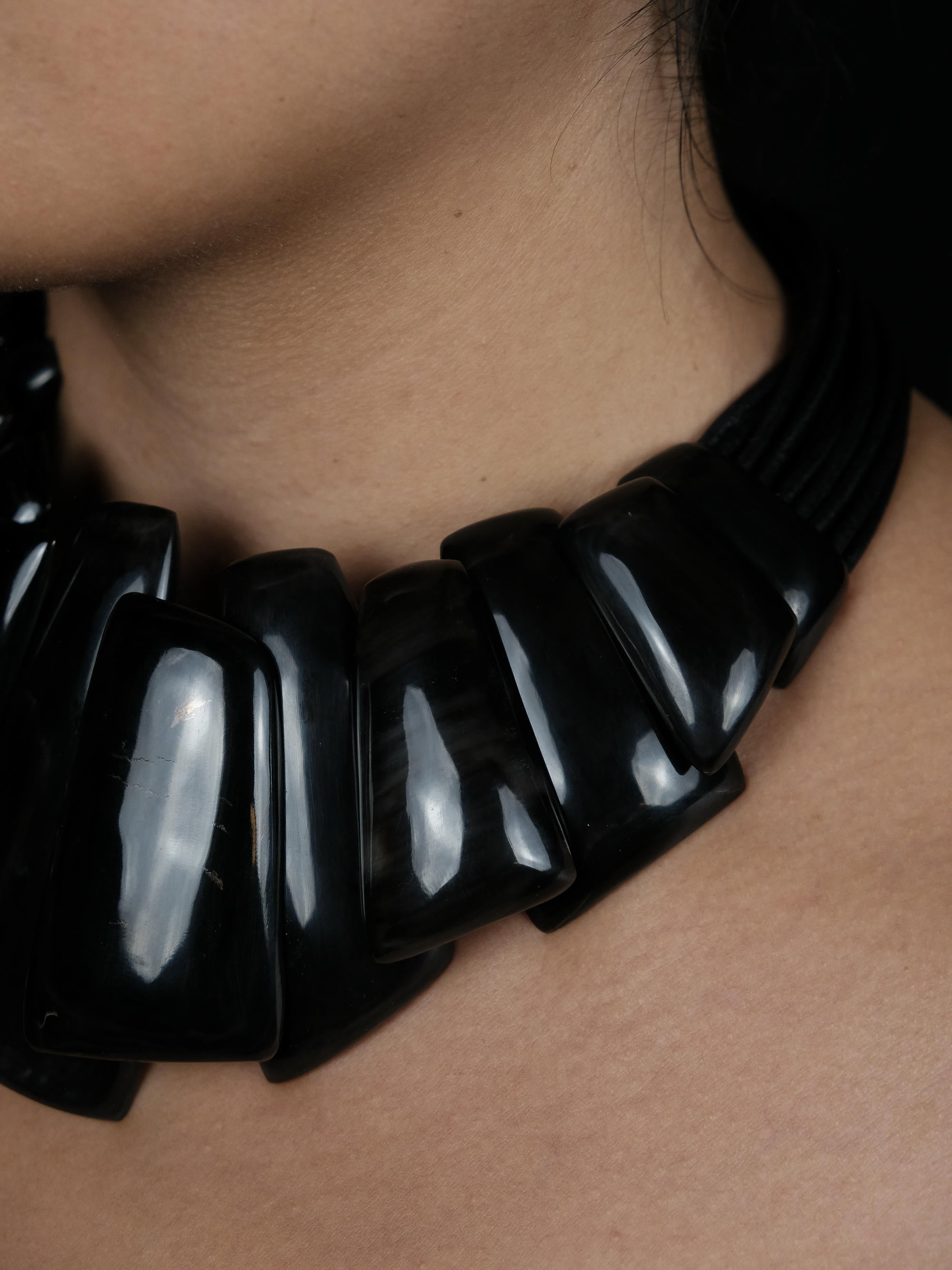 A close-up portrait of a woman wearing a chunky, glossy black collar necklace against a solid black background.