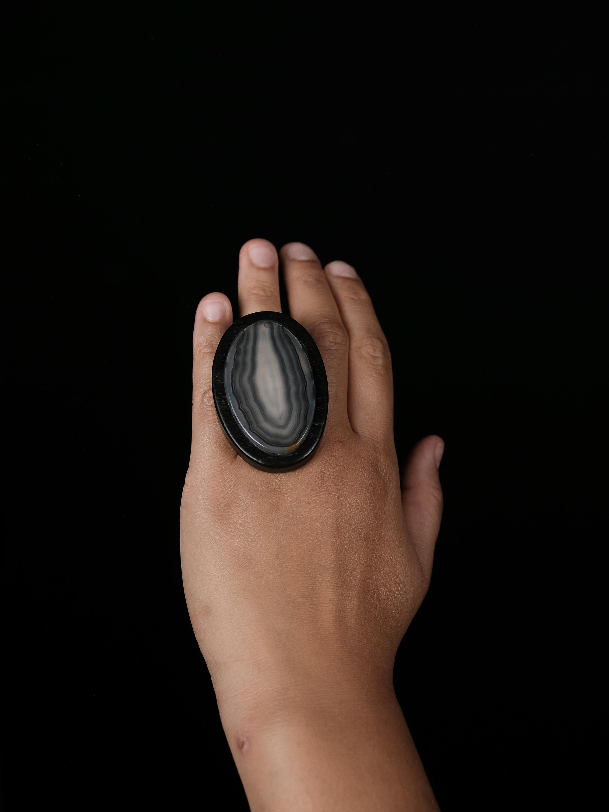 A hand wearing a large oval ring made from polished black agate with natural white and gray banding, set against a solid black background.