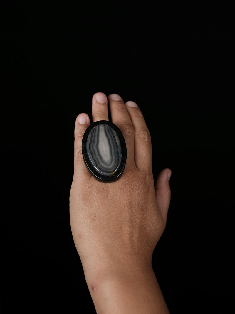 A hand wearing a large oval ring made from polished black agate with natural white and gray banding, set against a solid black background.