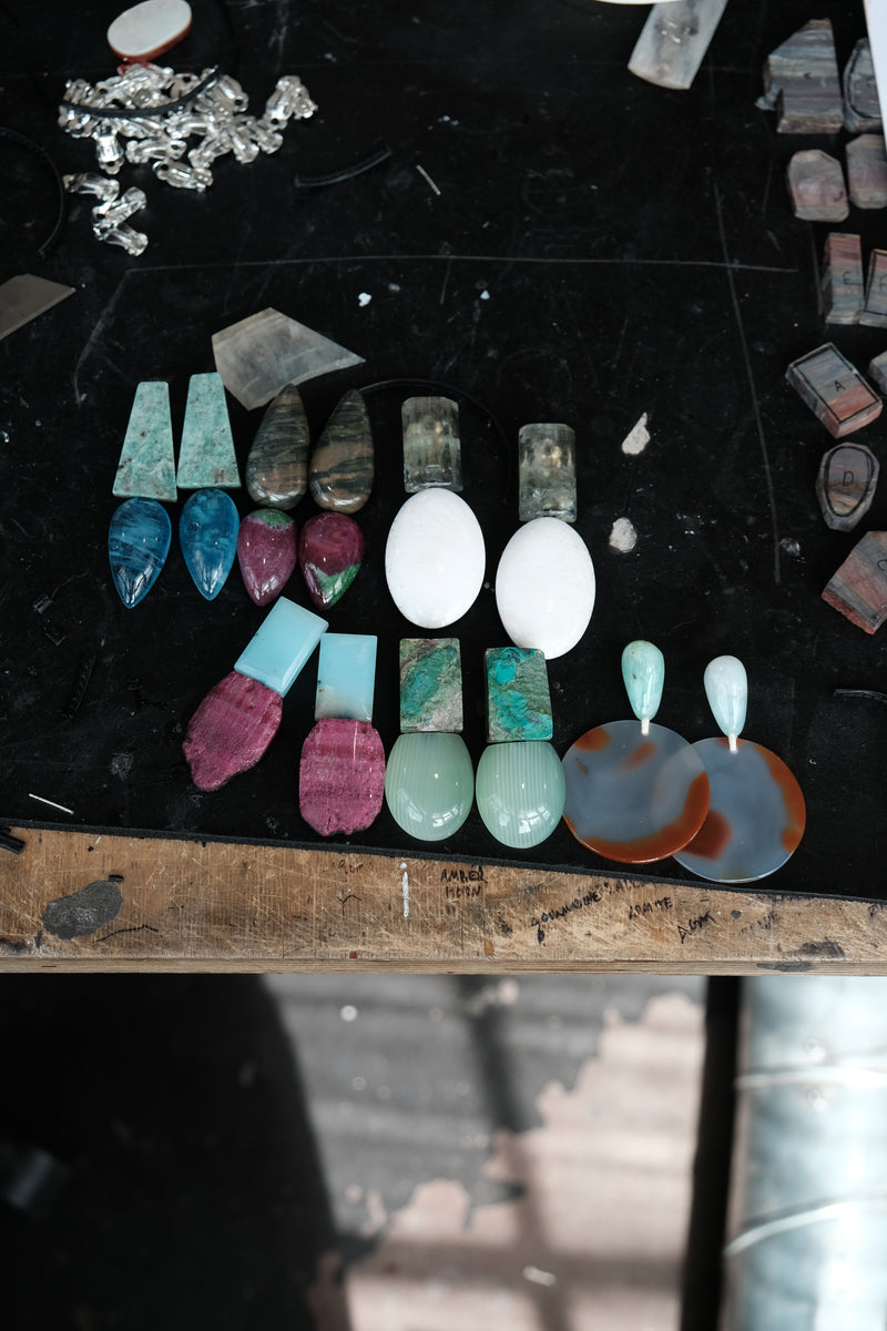 A collection of polished gemstone jewelry pieces, including earrings and pendants, arranged on a dark workbench. The stones feature various colors and patterns such as turquoise, white, pinkish-red, green, and marbled gray and orange.